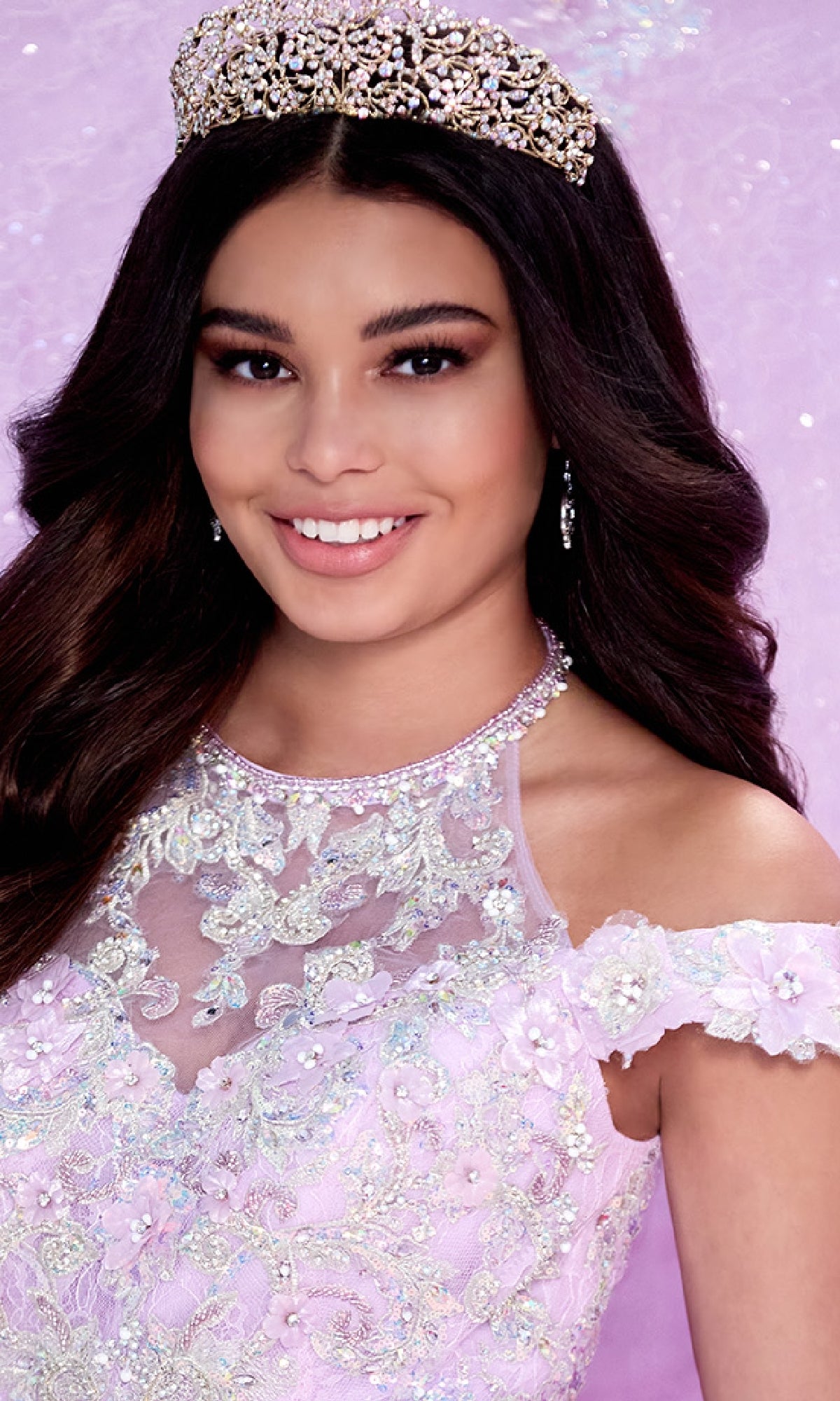A young woman with long dark hair smiles against a light purple background, wearing the jeweled tiara and ornate, off-the-shoulder lavender Quinceanera Dress PR30089 By Princesa, adorned with embroidered lace and beadwork.